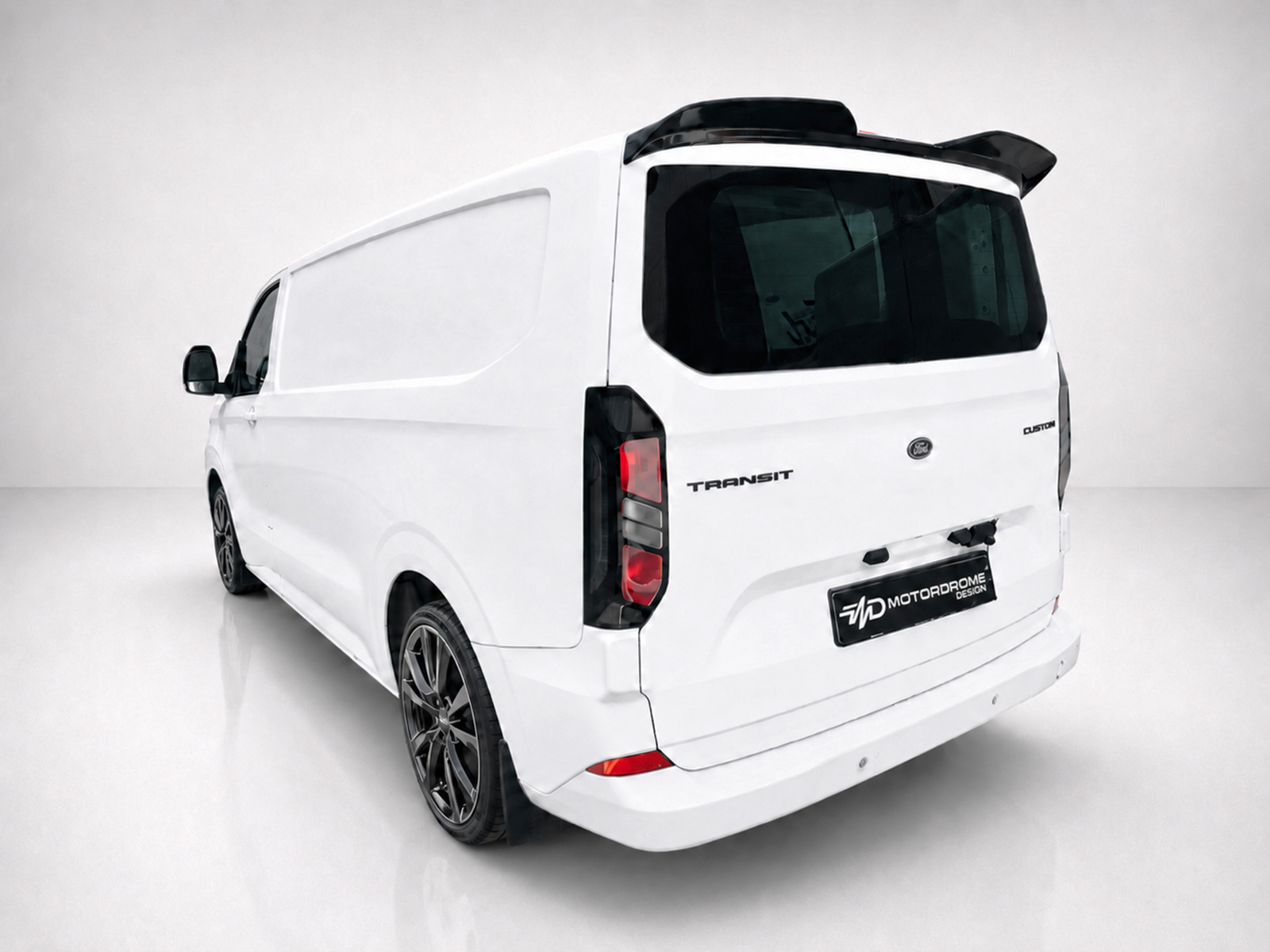 A-550_2jpg - OPSHOLDERS Rear three-quarter view of a white Ford Transit van with dark-tinted rear window and aftermarket black wheels; a custom logo plate reads 'MOTOR DROME DESIGN'.