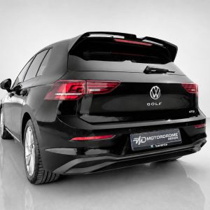 Rear view of a black Volkswagen Golf SUV with a roof spoiler in a bright studio setting, showing taillights and VW logo.",