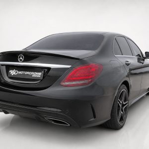 Rear three-quarter view of a black Mercedes-Benz sedan with tinted windows on a white studio backdrop, showing the trunk spoiler and chrome trim at the rear plate area.