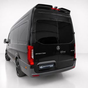 Rear three-quarter view of a black Mercedes-Benz Sprinter van in a bright studio, showing the tail lights, rear doors, and a Motordrome Design plate on the bumper.