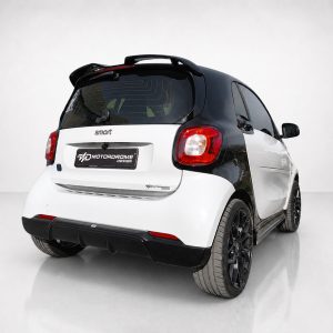 Rear three-quarter view of a two-tone black-and-white Smart car, showing red rear lights and a showroom-like background with Motordrome Design branding on the plate.