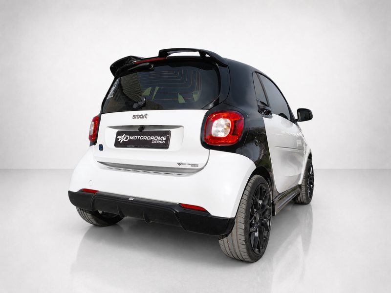 Rear three-quarter view of a two-tone black-and-white Smart car, showing red rear lights and a showroom-like background with Motordrome Design branding on the plate.