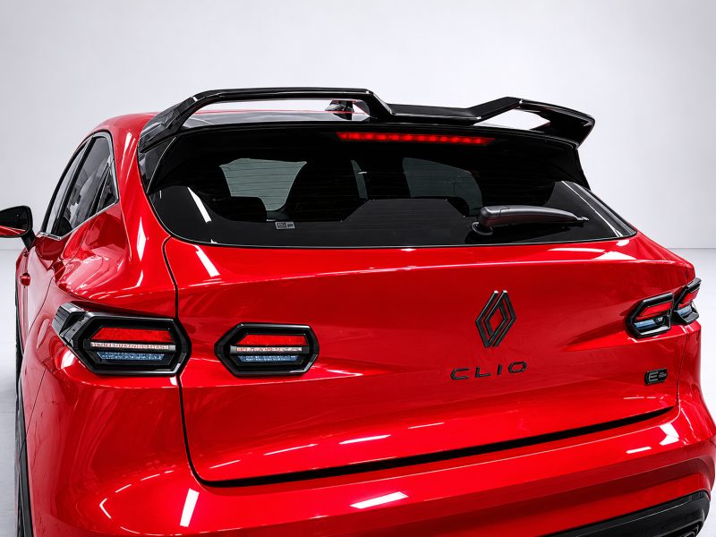 Rear view of a bright red Renault Clio with a black roof spoiler and distinctive split tail lights, showing the Renault badge and model name on the hatch.