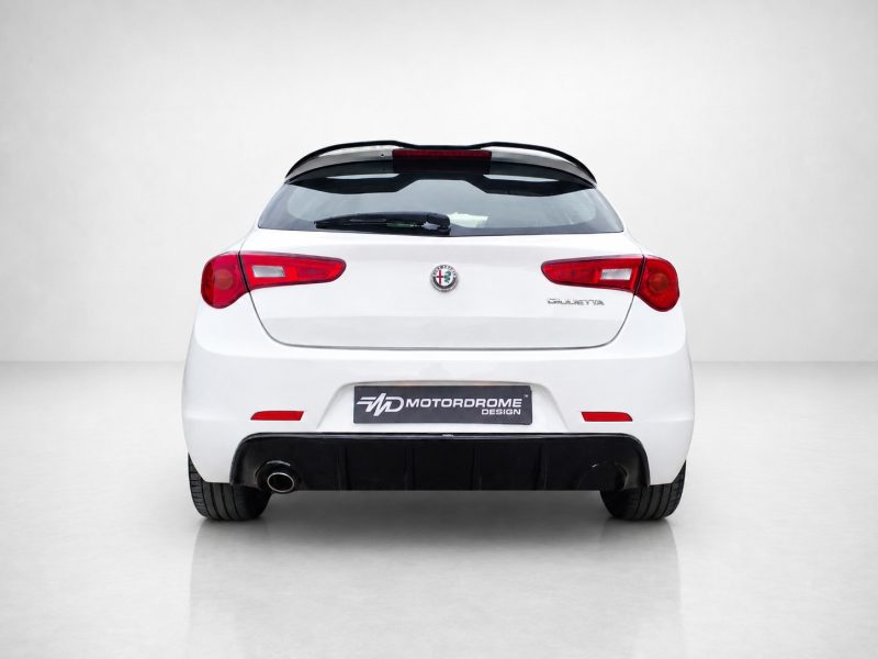 Rear view of a white Alfa Romeo Giulietta in a studio, showing tail lights, hatch, and a Motordrome Design plate above the bumper.