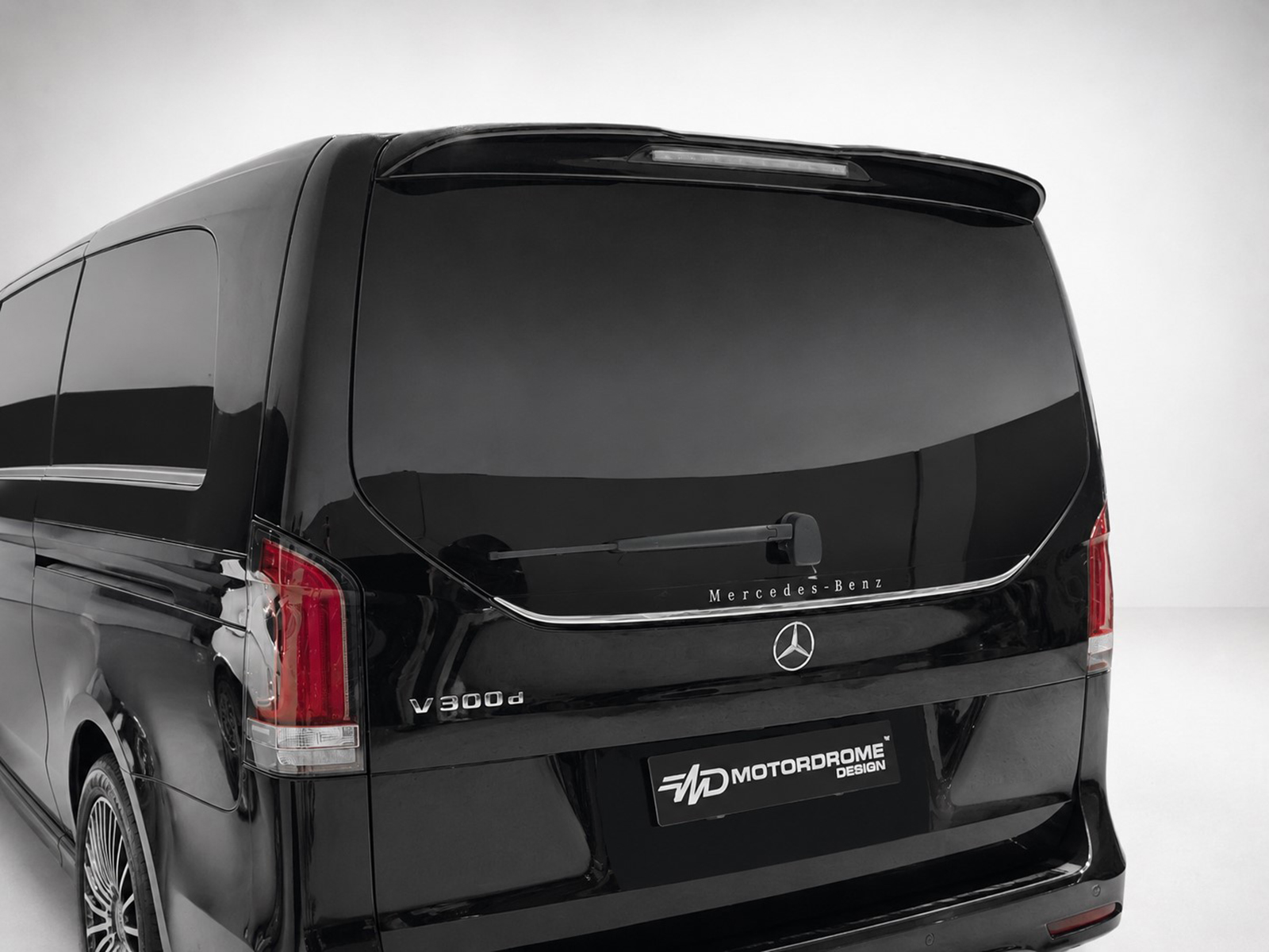 A-578_1jpg - OPSHOLDERS Rear view of a black Mercedes‑Benz V300d van with dark tinted windows and a MotorDrome Design plate on the bumper edge to the right of the emblem.