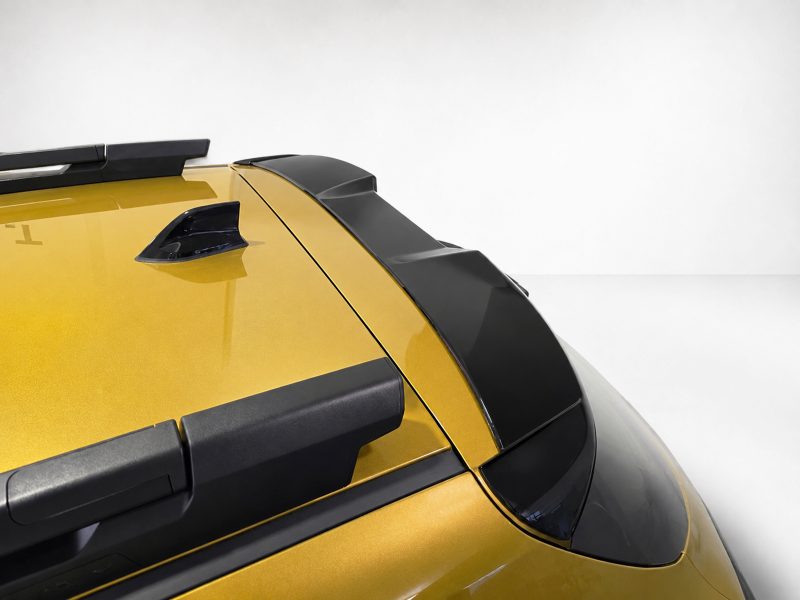 A-579_4jpg - OPSHOLDERS Close-up of the rear roof area of a yellow car, showing a roof rack, black spoiler, and antenna fin against a light background.