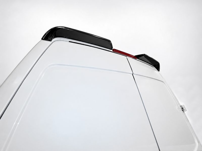 White van rear with black roof spoiler and slim red brake light strip against a pale sky.