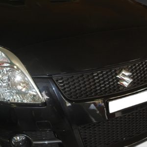 Front view of a black Suzuki car showing the grille, emblem, and headlight cluster on the left side of the image while parked indoors/at night.