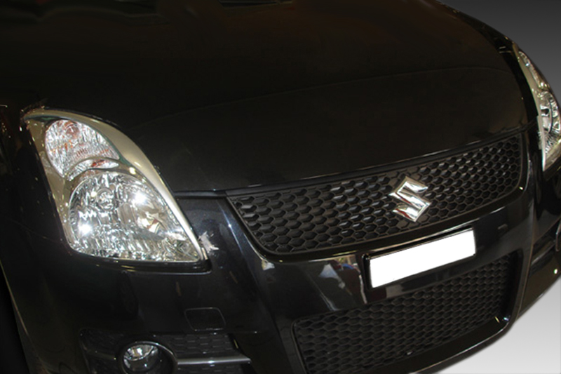 FR000104_1jpg - OPSHOLDERS Front view of a black Suzuki car showing the grille, emblem, and headlight cluster on the left side of the image while parked indoors/at night.