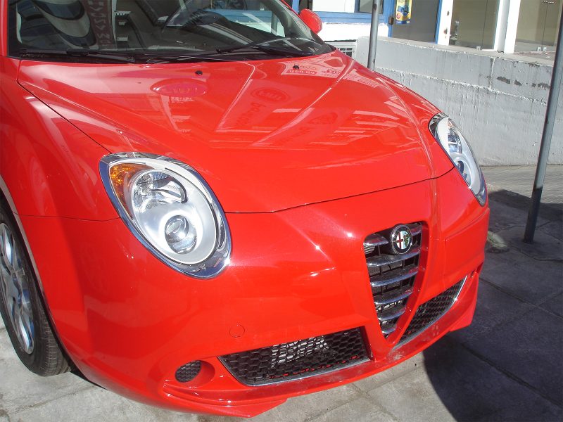 Front view of a bright red Alfa Romeo car, showing its round headlights and distinctive grille outdoors.