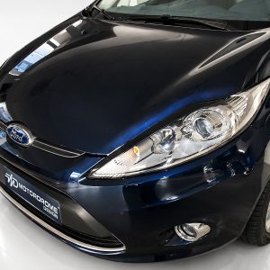 Close-up of a dark blue Ford car front, showing the hood, headlight, and emblem in a showroom setting