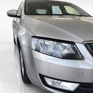 Close-up of the front-left side of a silver sedan, showing the headlight, grill, and hood in a bright showroom setting.