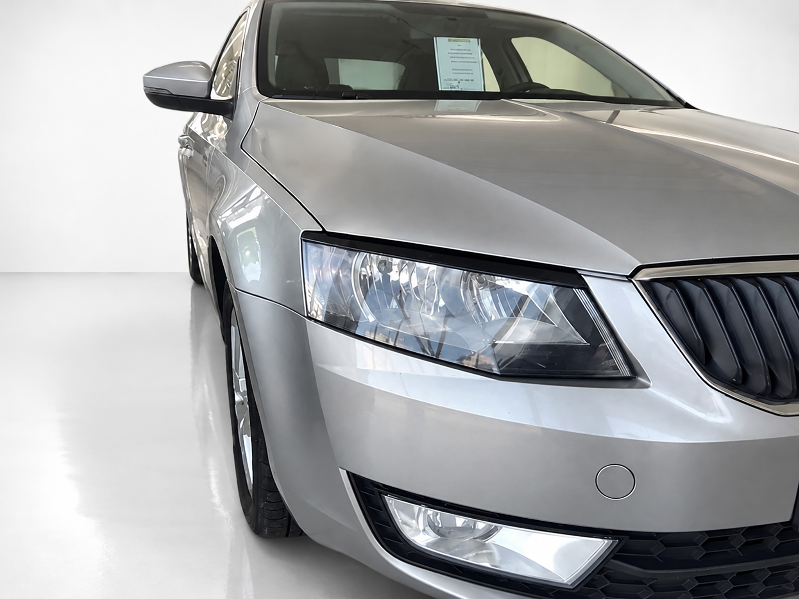 FR000146_1jpg - OPSHOLDERS Close-up of the front-left side of a silver sedan, showing the headlight, grill, and hood in a bright showroom setting.