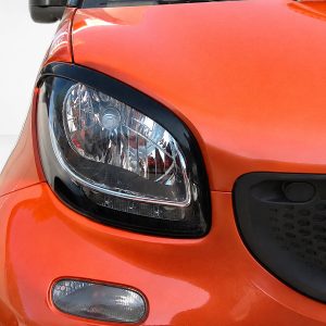 Close-up of an orange car's headlight and front body, showing the surrounding black trim and grille area on the right side of the image.