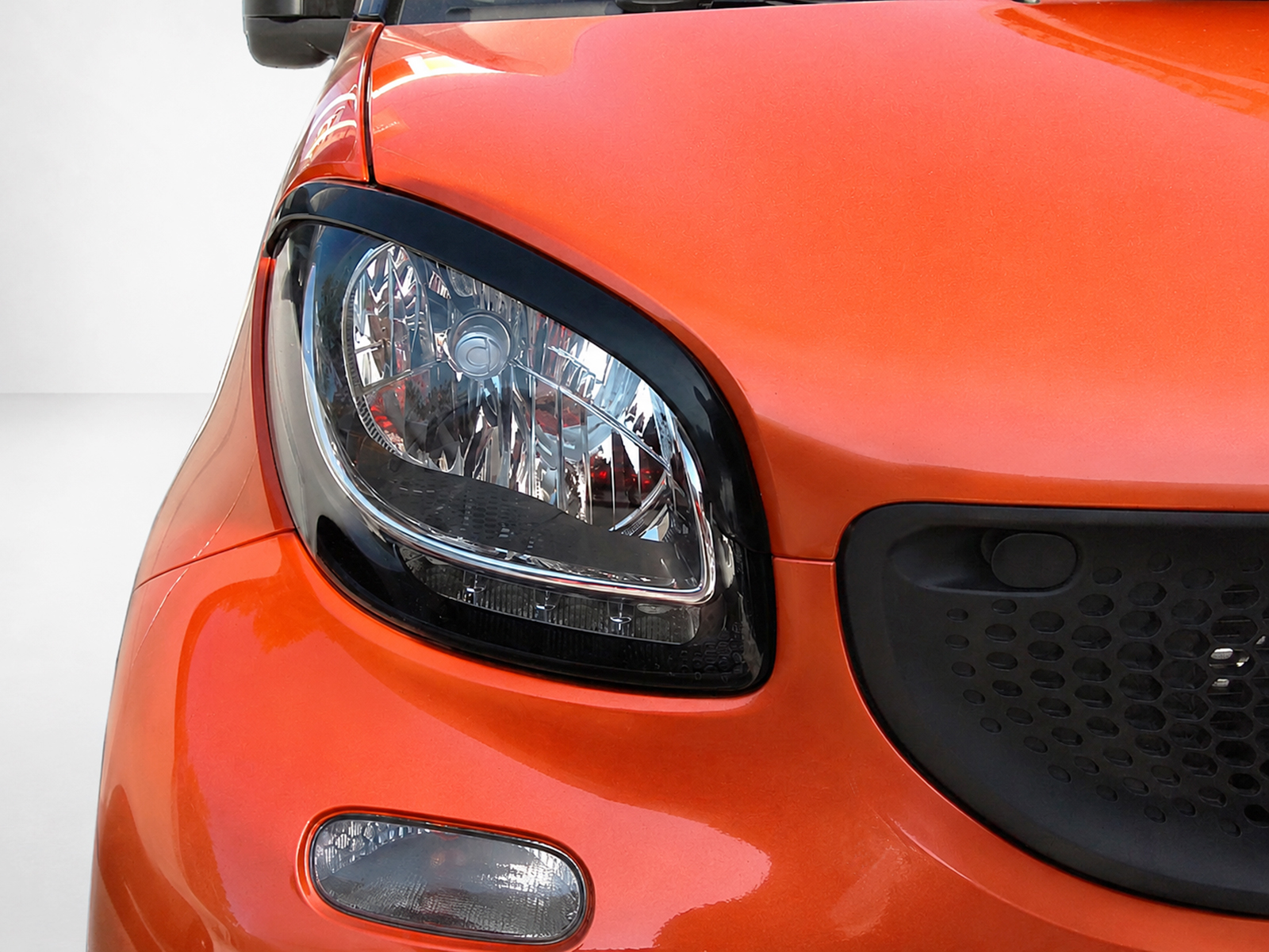 FR000153_1jpg - OPSHOLDERS Close-up of an orange car's headlight and front body, showing the surrounding black trim and grille area on the right side of the image.