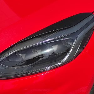 Close-up of a red car's front left area, showing the headlight, black trim, and part of the wheel.