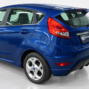 Rear three-quarter view of a blue Ford hatchback, showing the red taillight, rear window, and alloy wheel in a studio setting.