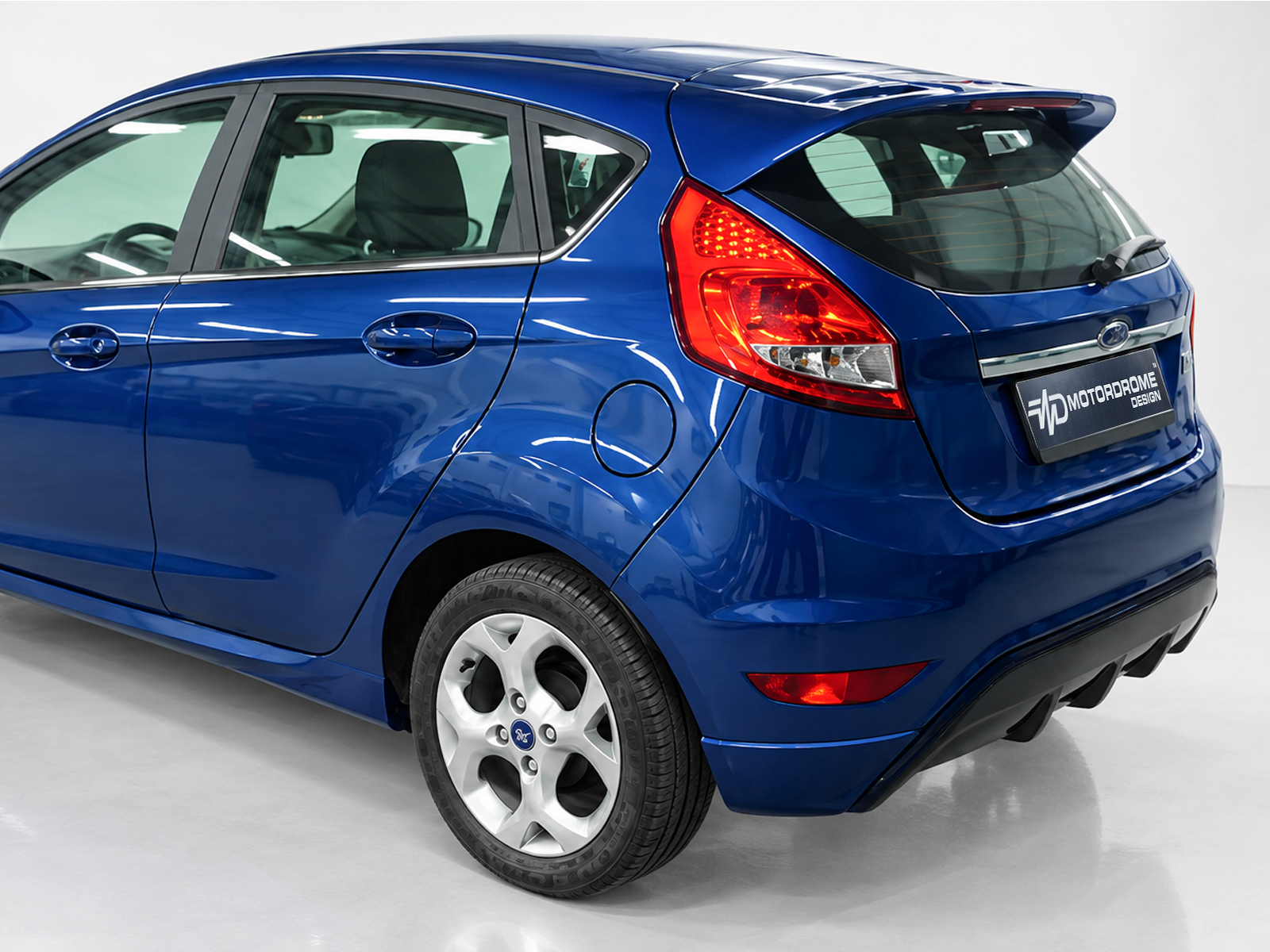 K124-006_1jpg - OPSHOLDERS Rear three-quarter view of a blue Ford hatchback, showing the red taillight, rear window, and alloy wheel in a studio setting.