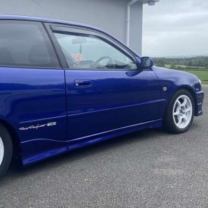 Blue sporty car photographed from the side, white alloy wheels, TRD badge on the lower door, parked on a paved area with a countryside background