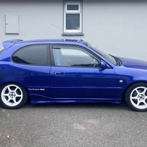 K126-003_2jpg - OPSHOLDERS Side view of a blue hatchback car with white aftermarket wheels parked on a street in front of a light gray wall.