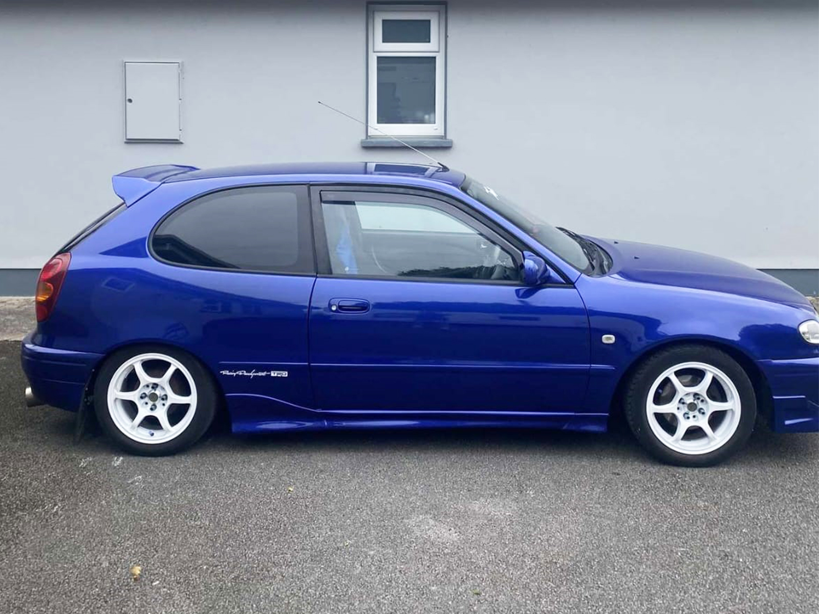 K126-003_2jpg - OPSHOLDERS Side view of a blue hatchback car with white aftermarket wheels parked on a street in front of a light gray wall.
