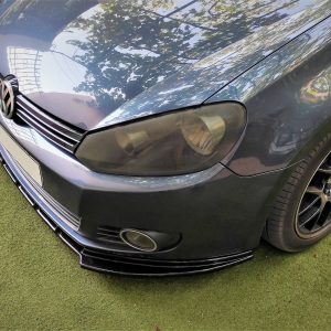 Front-left view of a dark VW car parked on green grass, showing headlight, grille, and alloy wheel with reflection on the hood.