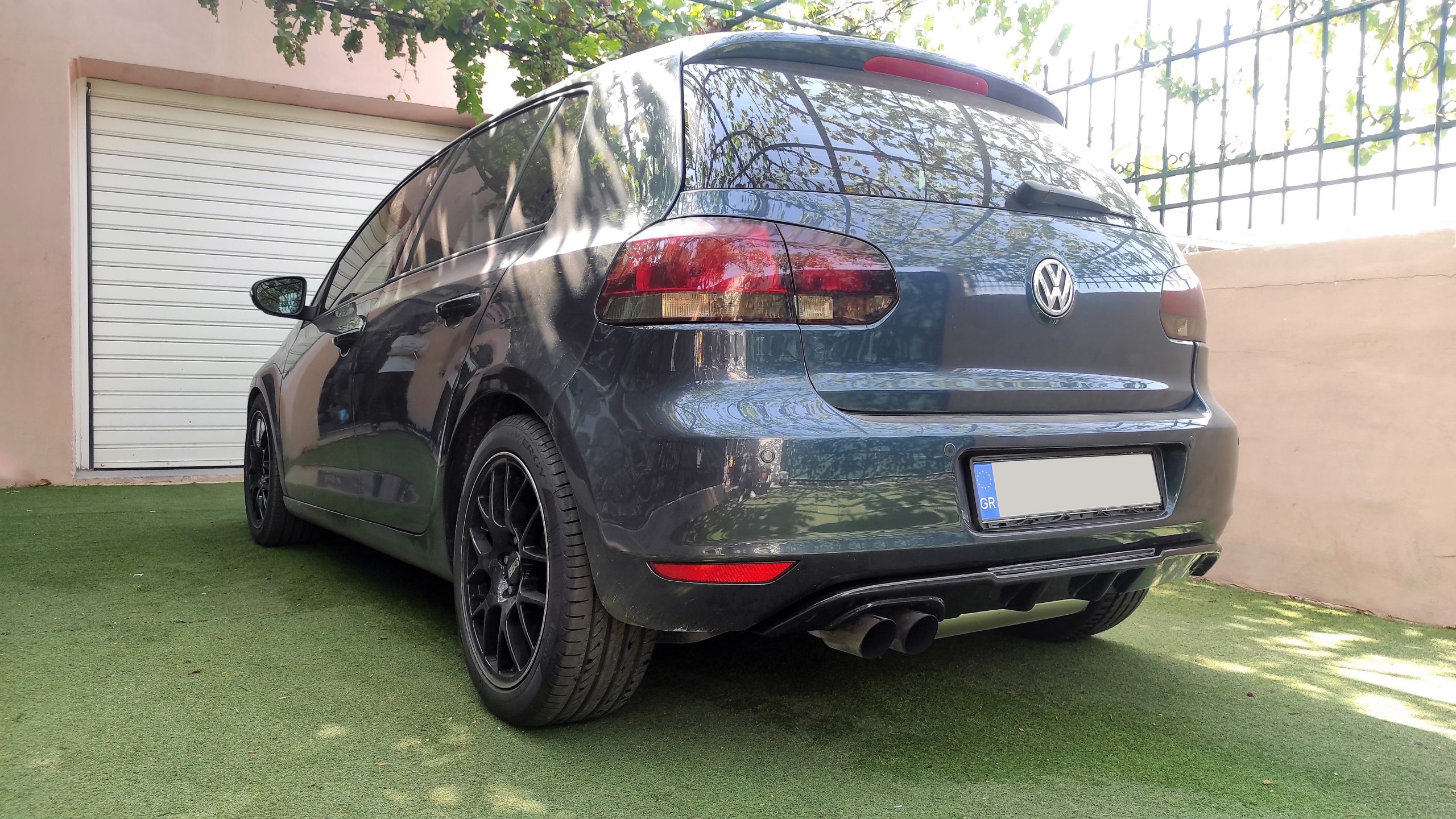 K128-006_2jpg - OPSHOLDERS Rear three-quarter view of a dark gray Volkswagen SUV parked on green artificial turf beside a beige wall and closed garage door, with reflections on the rear window.