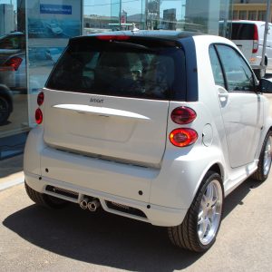 Rear three-quarter view of a white Smart car parked beside a glass storefront on a sunny day at a dealership lot.