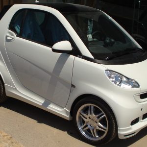 White compact car parked on a driveway, shown from the front-right side with alloy wheels and a black roof rail.