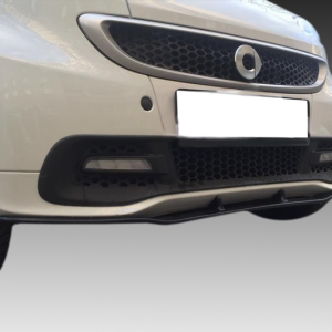 Close-up of a white car's front bumper, showing the black lower grille, fog light housings, and partially obscured license plate.