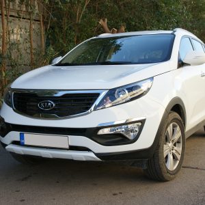 Front-left view of a white Kia SUV parked on a street, with green hedges and trees in the background and a blue GR license plate visible.