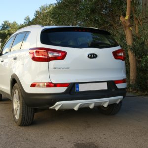 Rear view of a white Kia Sportage SUV parked on a paved lot with trees on the right side, branded logo visible.