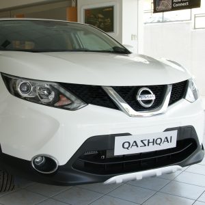 White Nissan Qashqai on a showroom floor, shown from the front-left with grille and headlight visible.