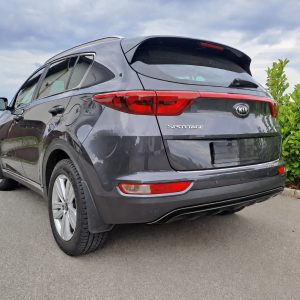 Gray Kia Sportage SUV parked on an asphalt lot beside green shrubs under a cloudy sky