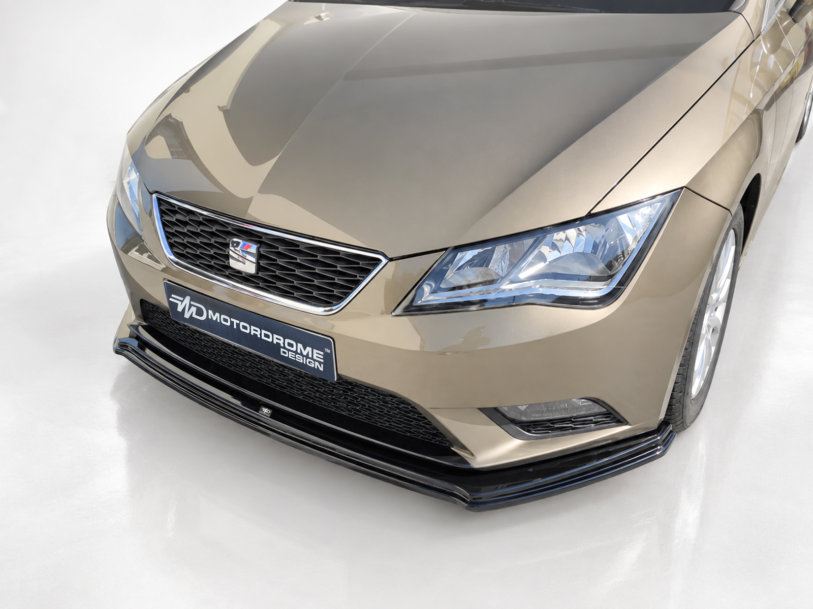 K150-007_2jpg - OPSHOLDERS Front three-quarter view of a metallic beige car facing left, showing a honeycomb grille with a small badge, sharp headlight, and a black lower front splitter with a motorsport-style plate reading MOTORDROME DESIGN