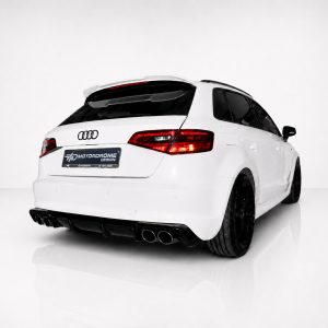 Rear three-quarter view of a white Audi with black wheels and quad exhausts in a studio setting, emphasizing the car's sporty rear end.