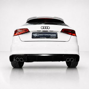 White Audi rear view in a studio, showing red taillights, quad exhausts, and a dark dealership plate reading MOTORDROME DESIGN (Gratsias).
