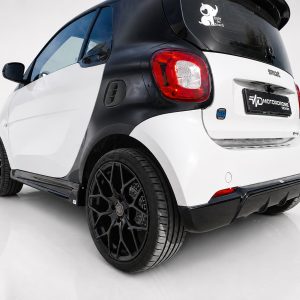 Rear three-quarter view of a white and black Smart car with oversized black wheels in a studio setting.