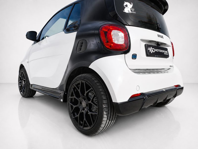 Rear three-quarter view of a white and black Smart car with oversized black wheels in a studio setting.