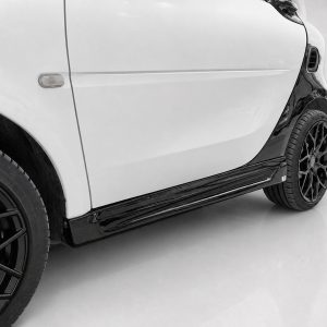 K154-003_3jpg - OPSHOLDERS Close-up of a white car's side, showing the door, small side marker light, and a black alloy wheel with a glossy lower side panel running along the bottom edge of the car.