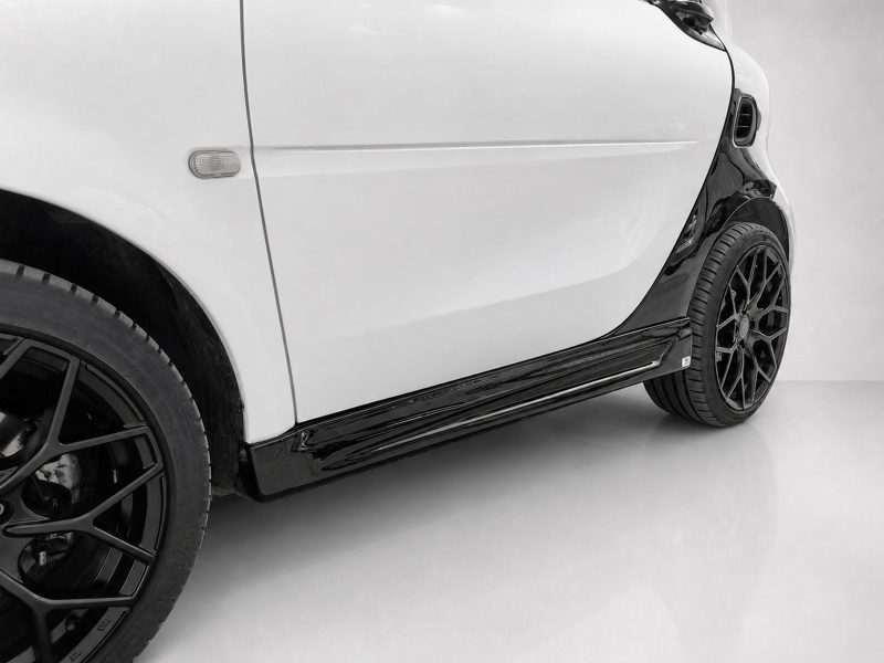 K154-003_3jpg - OPSHOLDERS Close-up of a white car's side, showing the door, small side marker light, and a black alloy wheel with a glossy lower side panel running along the bottom edge of the car.