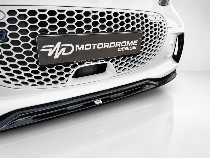K154-004_5jpg - OPSHOLDERS Front end of a white car with a hexagonal honeycomb grille and a Motordrome Design license plate showing the brand name Motordrome Design (logo area visible).