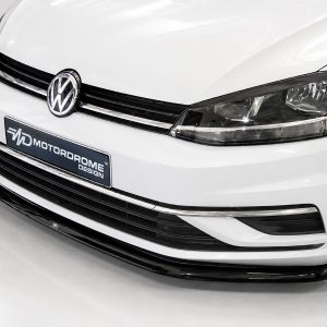 Close-up of a white VW car's front end, showing the grille, emblem, and headlight in a showroom setting