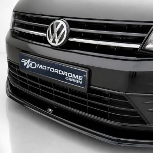 Close-up of a black Volkswagen front end with a chrome grille and centered VW emblem, featuring a bumper plate that reads MOTORDROME DESIGN