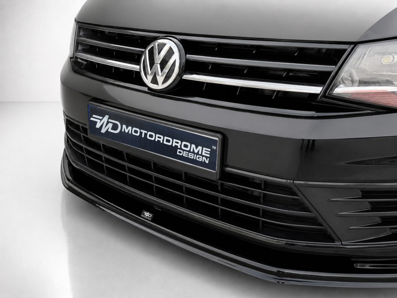 Close-up of a black Volkswagen front end with a chrome grille and centered VW emblem, featuring a bumper plate that reads MOTORDROME DESIGN