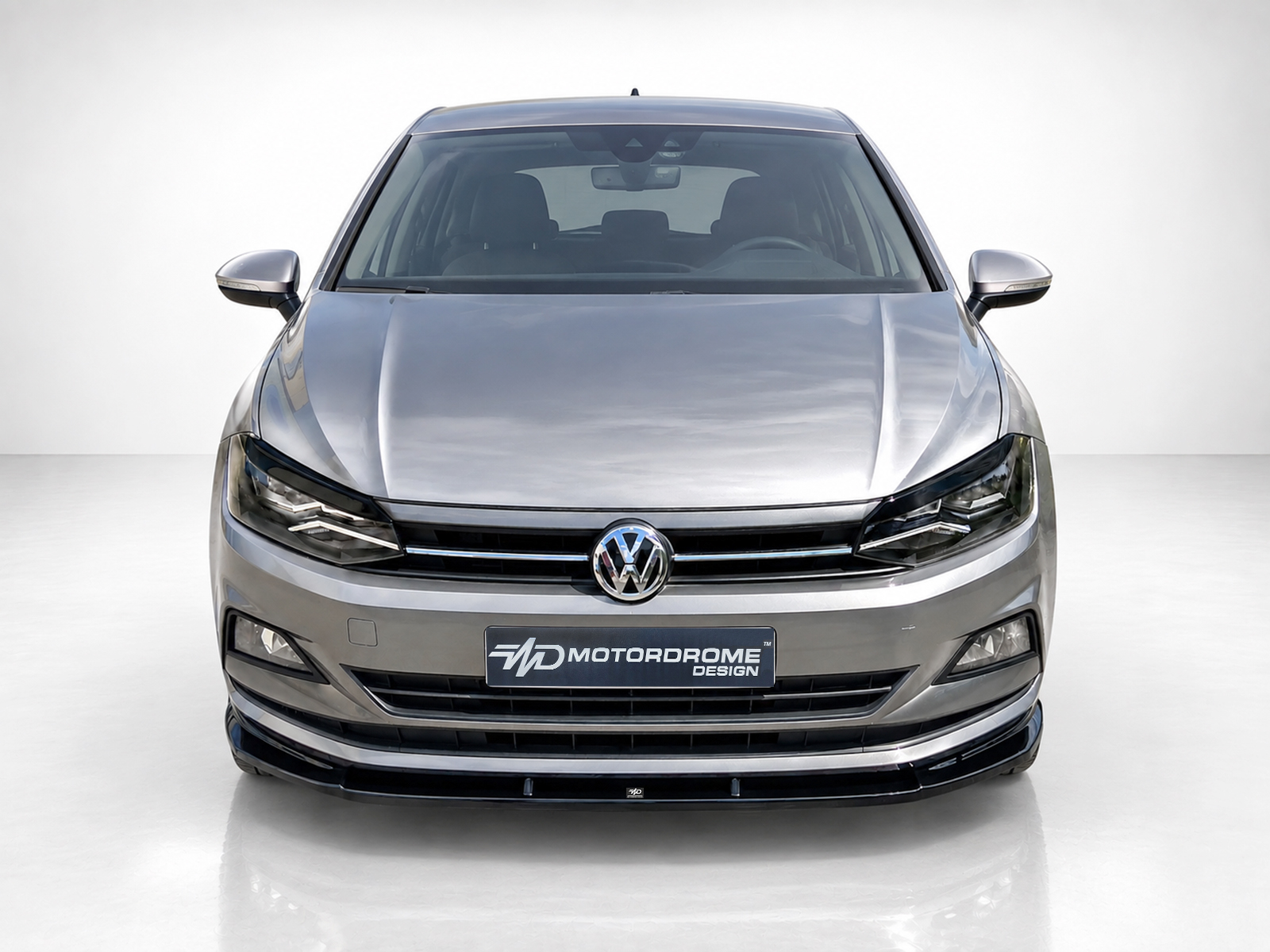 K162-001_1jpg - OPSHOLDERS Front view of a silver VW car facing the camera, showing the grille, logo, and headlights in a studio setting with a dealership plate reading 'Motordrome Design'