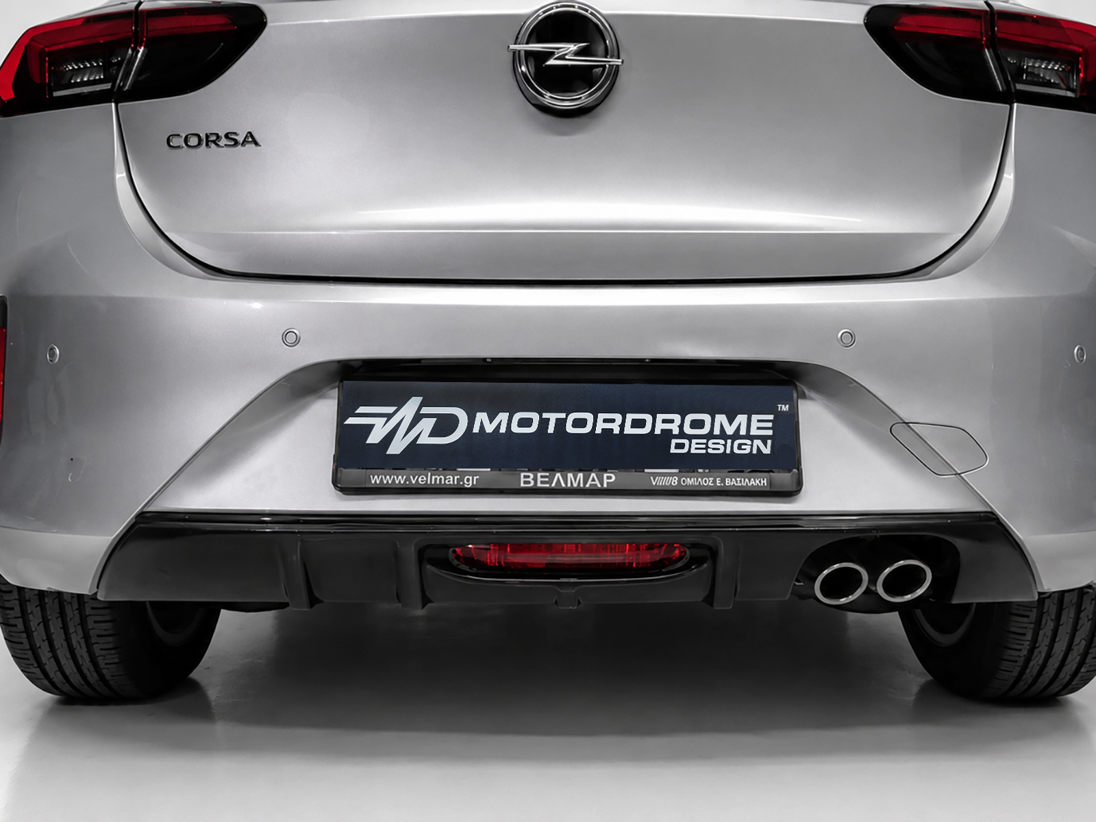 K163-004_1jpg - OPSHOLDERS Rear of a silver Opel Corsa with emblem, Corsa badge, red taillights, and a MOTORDROME DESIGN plate in a showroom setting, showing dual exhausts.