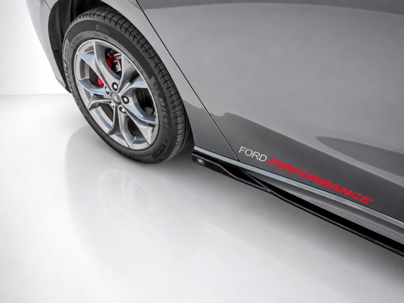 Close-up of a gray Ford car showing alloy wheel, red brake caliper, and the 'FORD PERFORMANCE' branding along the side skirt.