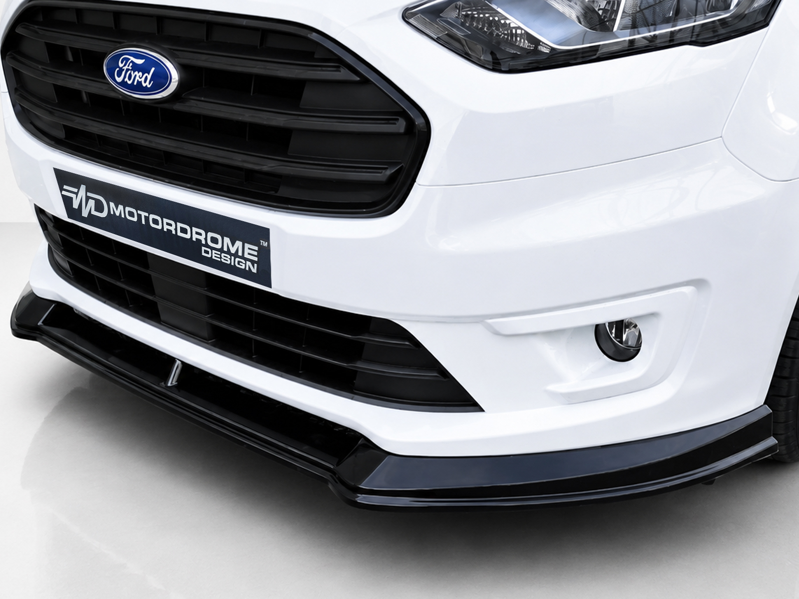 K167-001_2jpg - OPSHOLDERS Close-up of a white Ford car front, showing grille, headlight, and a MotorsDrome Design plate in the lower bumper area.