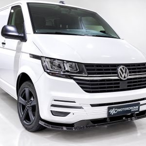 White Volkswagen Transporter van in a studio setting, front three-quarter view showing grille, headlights, and black alloy wheels with logo plate reading 'MOTORDROME'.